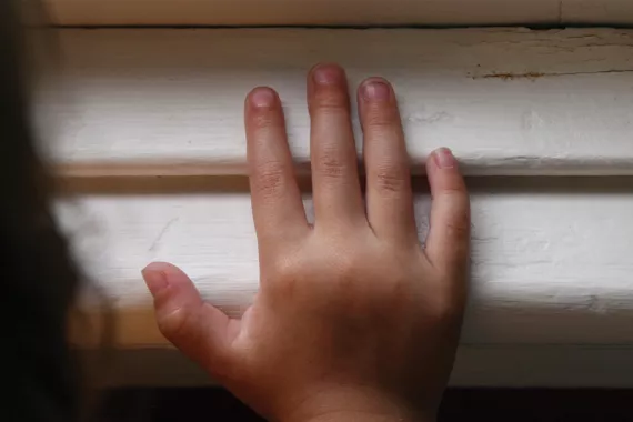 A child's hand is touching a horizontal window sill or wooden slats. The slats are painted solid white and appear slightly old and damaged