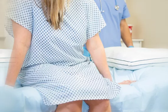 A woman wearing a hospital gown sits on a hospital bed. A man wearing scrubs and a stethoscope stands behind her. Their faces are not visible.