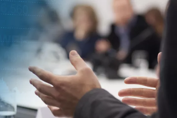 A pair of hands gestures to a group of people seen out of focus in the background. Stock market information is superimposed on the left.