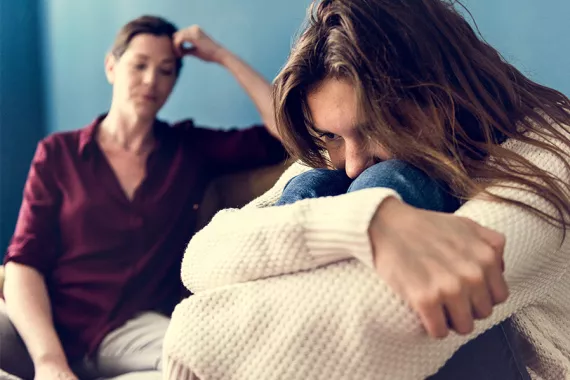 A teen girl sits hugging her knees and looking withdrawn and distressed. A woman in a red shirt sits behind her with a worried expression