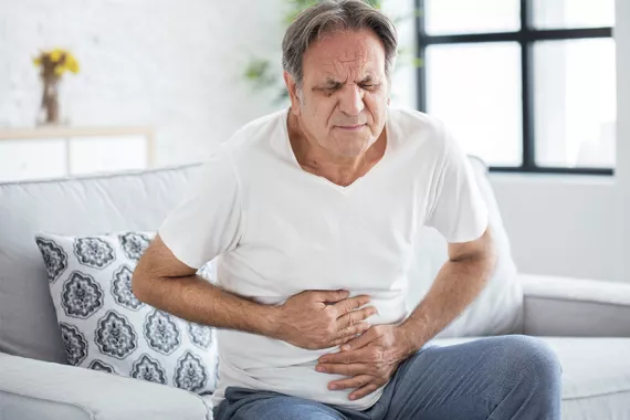 A middle-aged man is sitting on a couch with both hands covering his stomach. He looks like he is in pain.