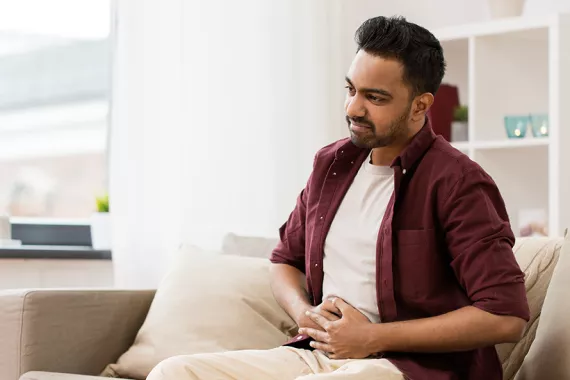 A man sits on a couch and holds his abdomen with a pained expression on his face